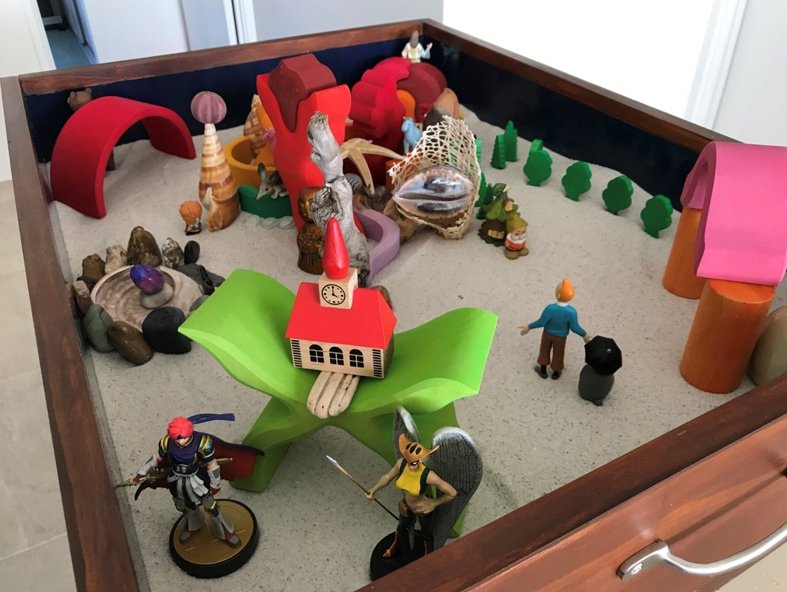 A Jungian Sandplay tray — wooden figures, stones, trees, a small church and seekers arranged in the sand